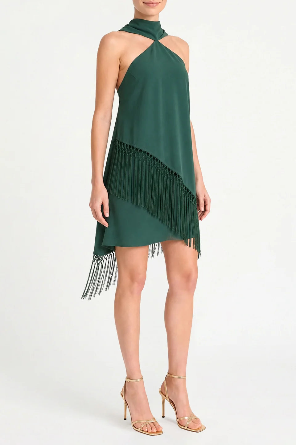 A woman modeling a dark green halter-neck mini dress with an asymmetrical fringe hem, paired with gold high-heeled sandals, against a plain white studio background.