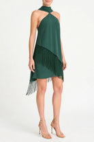A woman modeling a dark green halter-neck mini dress with an asymmetrical fringe hem, paired with gold high-heeled sandals, against a plain white studio background.
