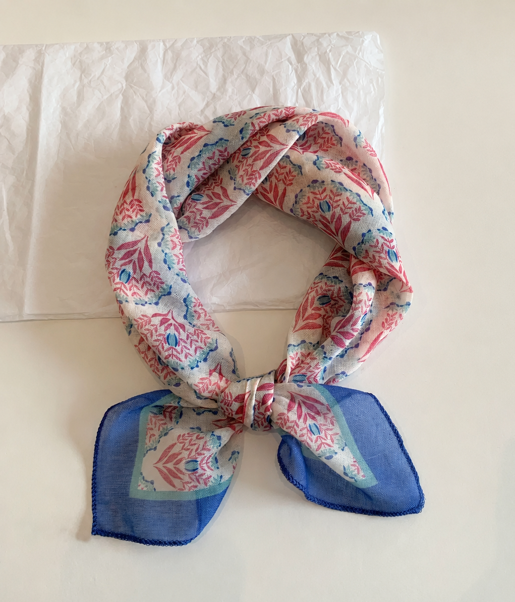 A square silk scarf with a pink, blue, and white floral pattern, tied in a knot and displayed on a light beige surface with a white tissue paper in the background.