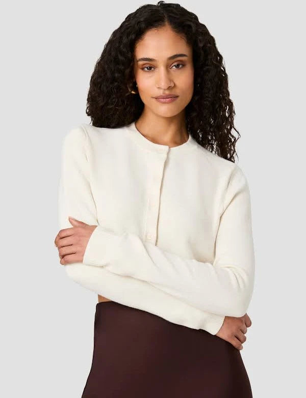 A woman with curly dark hair wearing a cream-colored long-sleeve knit cardigan with a buttoned front and a dark brown skirt, posed against a solid light gray background.