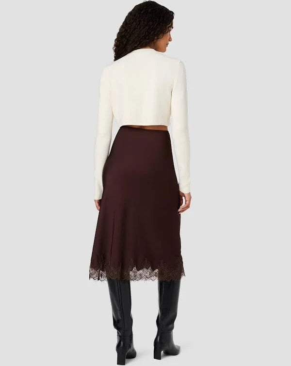 A woman shown from the back wearing a cream long-sleeve cropped cardigan and a dark brown midi skirt with a lace hem, paired with black knee-high boots, against a light gray background.