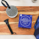 A boxed men's grooming product labeled 'Spuncalgie Men's Triple Butter Supreme Buffer' sits on a wooden board, alongside a black razor, a round scrubber, and part of a blue striped necktie. The product box is the main subject and is photographed from above on a wood surface.
