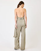 A woman shown from the back wearing a light taupe open-back jumpsuit with wide-leg trousers and a draped side detail, photographed against a plain light gray studio background.