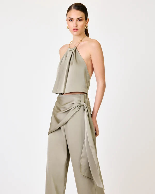 A model wearing a matching two-piece set in a light sage green satin fabric, featuring a halter crop top and high-waisted draped wide-leg pants, set against a plain white studio background.