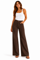 A woman modeling a pair of dark brown wide-leg pants with a white tank top, standing against a plain white background.
