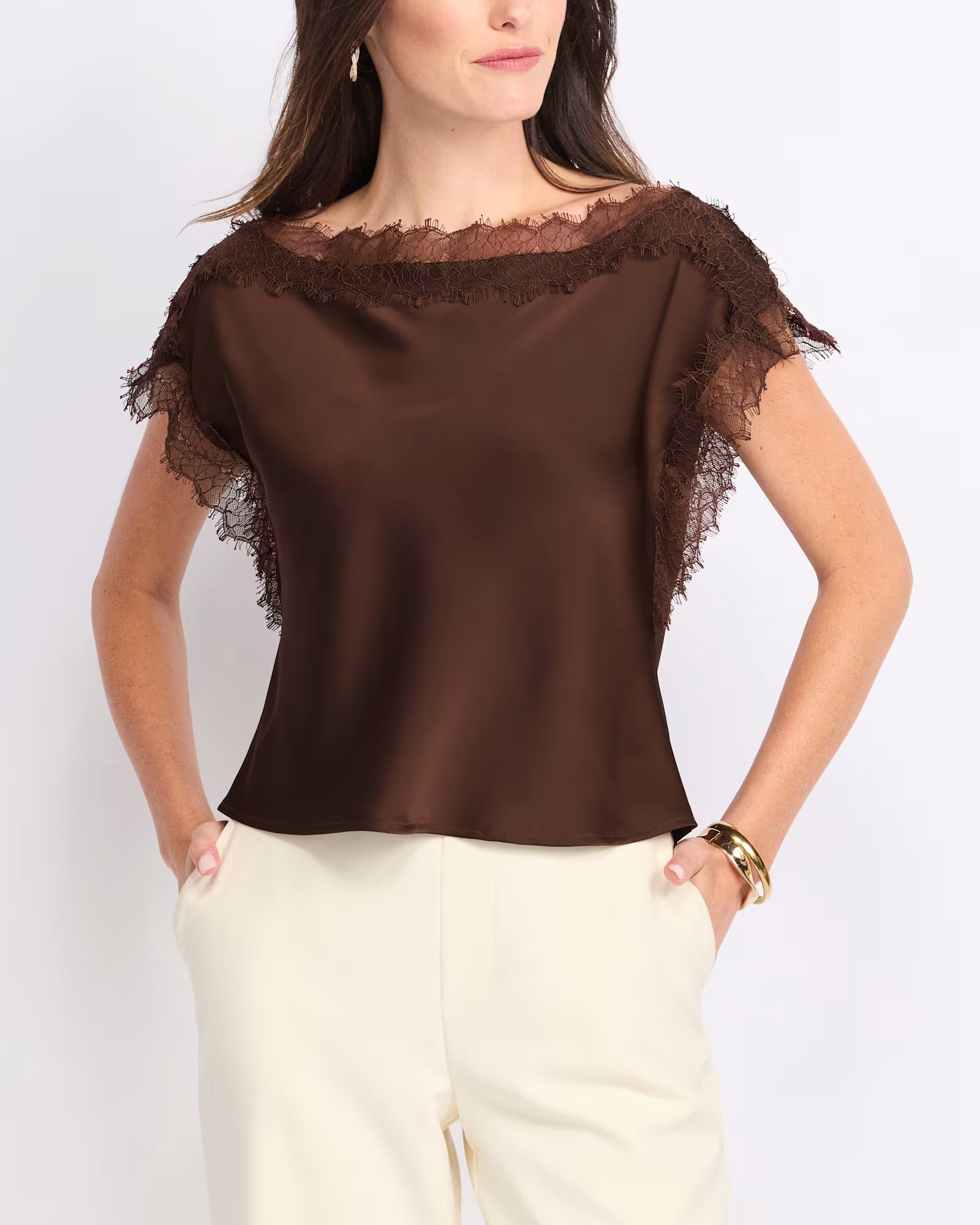 A woman wearing a brown satin short-sleeve top with sheer lace trim at the neckline and sleeves, paired with cream-colored pants, against a plain light background.