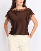A woman wearing a brown satin short-sleeve top with sheer lace trim at the neckline and sleeves, paired with cream-colored pants, against a plain light background.